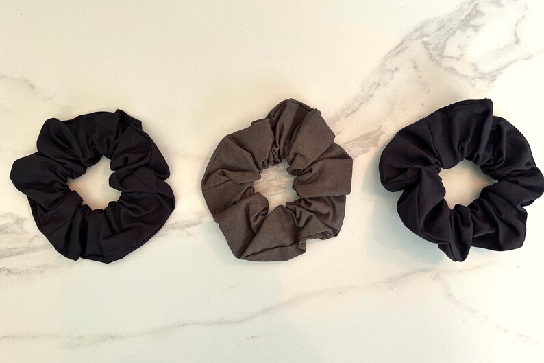 May include: Three fabric scrunchies in black, dark gray, and light gray. The scrunchies are made of soft, stretchy fabric and are perfect for holding hair in place.