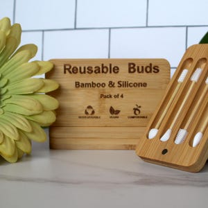Op de afbeelding: Herbruikbare oorstokjes van bamboe en siliconen. De houten doos is gegraveerd met "Reusable Buds Bamboo & Silicone Pack of 4". De stokjes zitten in een houten houder. Een gele bloem en een steen op de achtergrond.