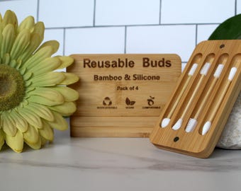 May include: A set of reusable bamboo and silicone ear buds. The wooden box is engraved with "Reusable Buds Bamboo & Silicone Pack of 4". The buds are in a wooden holder. A yellow flower and a rock are in the background.
