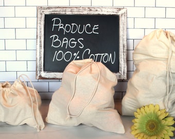 May include: Three white cotton produce bags with drawstring closures. The bags are sitting on a white tiled countertop. A chalkboard sign behind the bags reads "PRODUCE BAGS 100% COTTON".