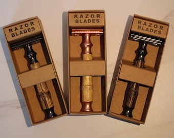 May include: Three safety razors in cardboard boxes with the text "RAZOR BLADES" on the front. The razors have wooden handles and different colored metal heads: black, copper, and silver.