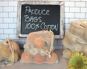 May include: Three reusable produce bags made of 100% cotton mesh, filled with oranges and lemons. The bags are tied with a drawstring and are displayed on a white tiled countertop. A chalkboard sign above the bags reads "PRODUCE BAGS 100% COTTON".