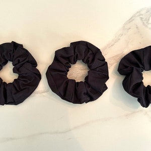 May include: Three black scrunchies made of soft fabric. The scrunchies are arranged in a row on a white marble surface.