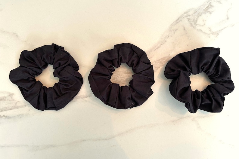 May include: Three black scrunchies made of soft fabric. The scrunchies are arranged in a row on a white marble surface.