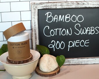 May include: A brown cardboard tube with a white lid containing 200 eco-friendly bamboo cotton swabs. The tube is sitting on a wooden coaster. A chalkboard sign reads "Bamboo Cotton Swabs 200 Piece".