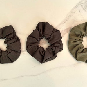May include: Three fabric scrunchies in different shades of brown and green. The scrunchies are made of soft, stretchy fabric and are perfect for holding hair in place.