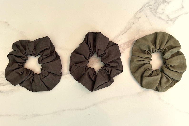 May include: Three fabric scrunchies in different shades of brown and green. The scrunchies are made of soft, stretchy fabric and are perfect for holding hair in place.