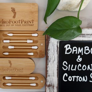 May include: Bamboo and silicone cotton swabs in bamboo cases. The cases are rectangular with rounded edges and feature the text "EnviroFootPrint" and a footprint logo. The image also includes a chalkboard sign that reads "Bamboo & Silicone Cotton Swabs."