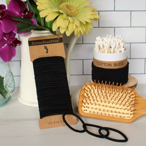 May include: A set of black hair ties, a wooden hair brush, and a container of cotton buds. The hair ties are packaged in a brown cardboard wrapper with the text "EnviroFootPrint" and "30 PIECES". The cotton buds are in a white container with a black band around the top that says "COTTON BUDS".
