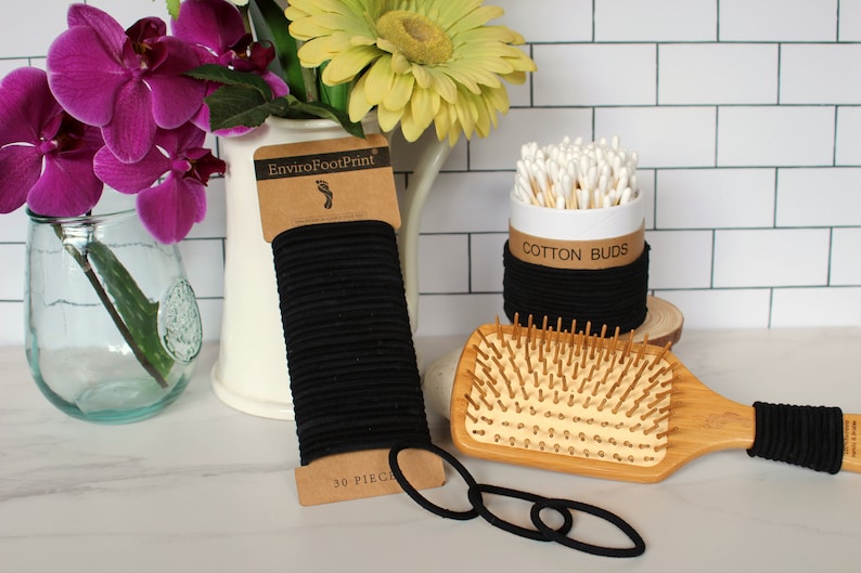 May include: A set of black hair ties, a wooden hair brush, and a container of cotton buds. The hair ties are packaged in a brown cardboard wrapper with the text "EnviroFootPrint" and "30 PIECES". The cotton buds are in a white container with a black band around the top that says "COTTON BUDS".