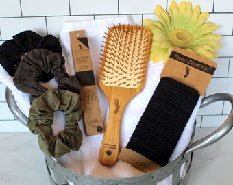May include: A white towel with a bamboo hair brush, a package of black hair ties labeled "EnviroFootPrint 30 PIECES", and a package of bamboo eyebrow razors labeled "Bamboo Eyebrow Razor". There are also three scrunchies in black, brown, and green.