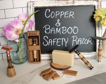 May include: A copper and bamboo safety razor with a wooden handle and a chrome blade. The razor is on a white countertop with a bar of soap, a wooden stand, and a package of razor blades.