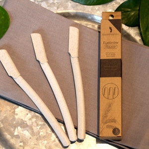 May include: Three white eyebrow razors with a brown cardboard box that says "Eyebrow Razor" and has a wheat stalk graphic. The box also says "Eco-friendly,  Disposable,  Derma safe".