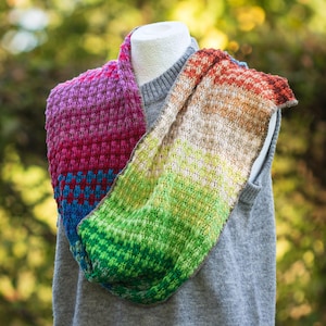 Puede incluir: Una bufanda infinita de punto multicolor con un patrón texturizado. La bufanda hace la transición del morado al rojo, azul, verde y marrón. La bufanda está colocada sobre un suéter gris en una cabeza de maniquí blanca.