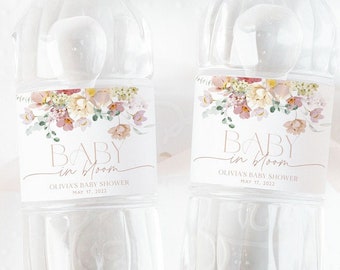 Può includere: Due bottiglie d'acqua in plastica trasparente con etichette floreali che recitano "Baby in Bloom" in uno schema di colori rosa e oro. Le etichette includono anche il testo "Olivia's Baby Shower" e "May 17, 2022".