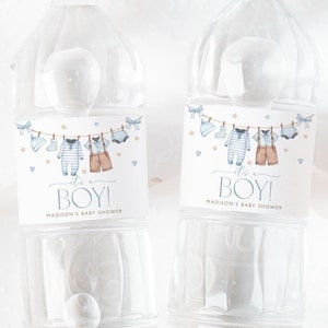 Op de afbeelding: Twee doorzichtige plastic waterflessen met blauwe en gouden etiketten. De etiketten tonen een rij babykleding die aan een waslijn hangt met sterren en harten. De tekst op de etiketten luidt "Het is een jongen!" en "Baby shower van Madison".