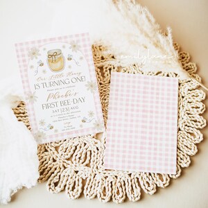 May include: Pink gingham patterned invitation with the text "Our Little Honey IS TURNING ONE!" and "Phoebe's FIRST BEE-DAY". The invitation features a honey pot, bees, and daisies. A blank pink gingham card is also present.