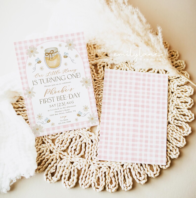 May include: Pink gingham patterned invitation with the text "Our Little Honey IS TURNING ONE!" and "Phoebe's FIRST BEE-DAY". The invitation features a honey pot, bees, and daisies. A blank pink gingham card is also present.