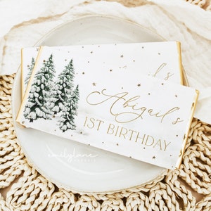 May include: White chocolate bar wrapper with a winter scene featuring three snow-covered pine trees. The text "Abigail's 1st Birthday" is in gold lettering.