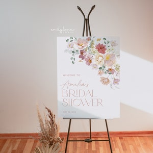 May include: A white welcome sign with a floral design in shades of pink, purple, and green. The text reads "Welcome to Amelia's Bridal Shower May 14, 2022".