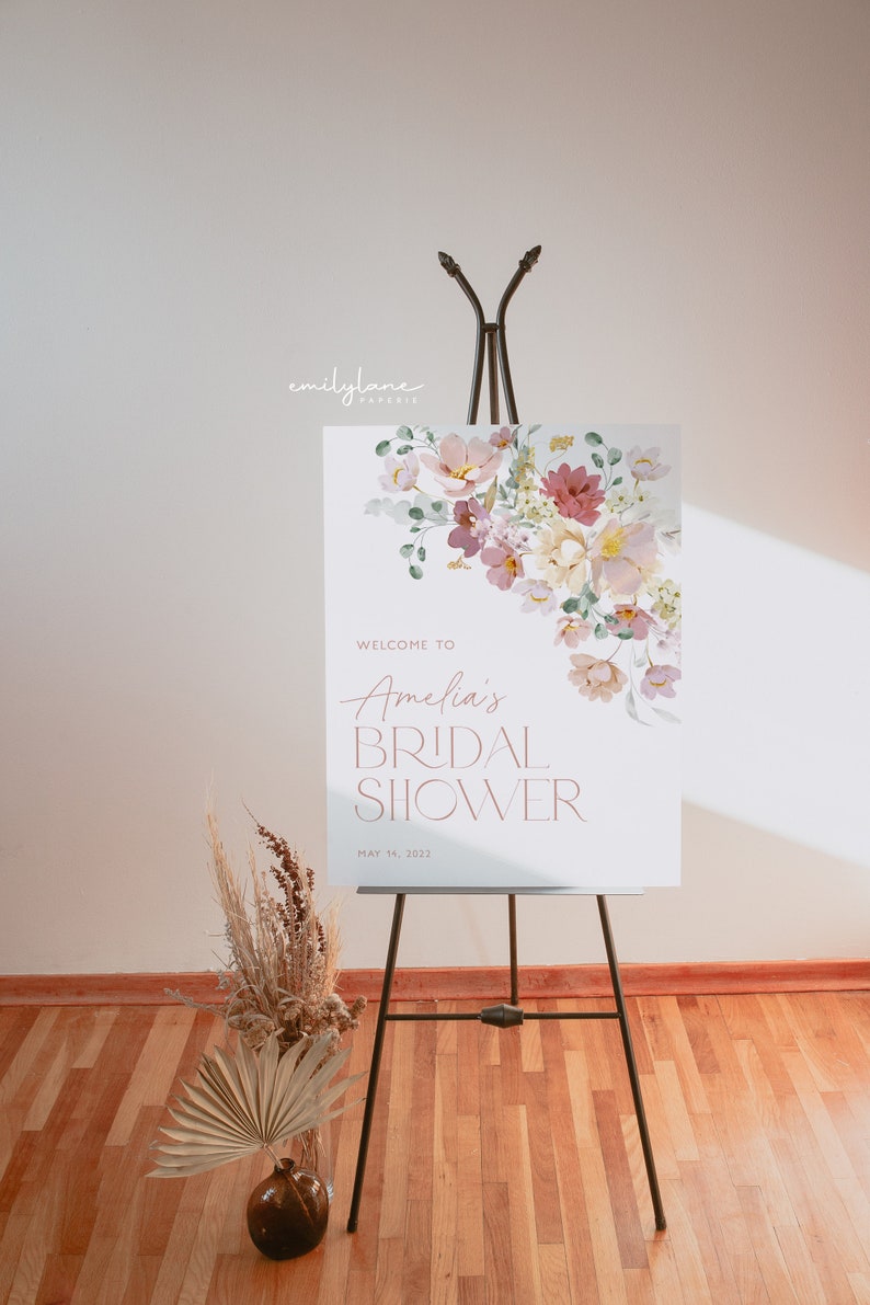 May include: A white welcome sign with a floral design in shades of pink, purple, and green. The text reads "Welcome to Amelia's Bridal Shower May 14, 2022".