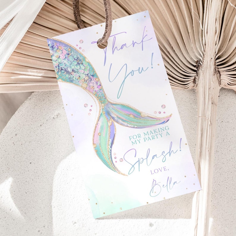 Puede incluir: Una etiqueta de agradecimiento con una ilustraci&oacute;n de cola de sirena. La cola es verde, morada y dorada con detalles de purpurina. El texto dice "&iexcl;Gracias! Por hacer de mi fiesta un Splash! Con cari&ntilde;o, Bella."