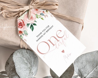Puede incluir: Una etiqueta de regalo blanca con detalles florales y las palabras "Thank you for making my birthday" y "One derful" en purpurina oro rosa. También hay un pequeño corazón y una firma.