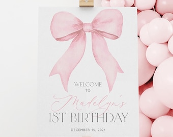 May include: A white sign on a wooden easel with a large pink watercolor bow illustration. The sign reads "WELCOME TO Madelyn's 1ST BIRTHDAY" and "DECEMBER 14, 2024" in pink and black text. Pink balloons are in the background.
