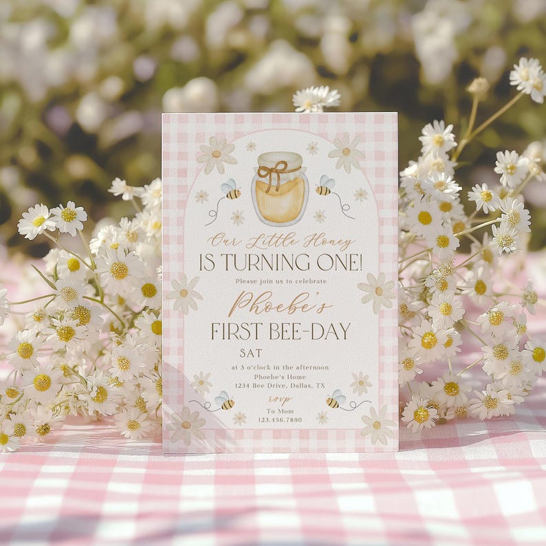 May include: A pink and white gingham patterned invitation with the text "Our Little Honey Is Turning One!" and "Phoebe's First Bee-Day". The invitation features a honey jar, bees, and daisy illustrations. The invitation is surrounded by white flowers.