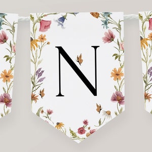 K&ouml;nnte beinhalten: Ein wei&szlig;es Banner mit einem floralen Rand und dem Buchstaben "N" in Schwarz in der Mitte. Die Blumen sind in den Farben Rosa, Gelb, Orange und Lila. Es gibt auch kleine Schmetterlinge im Design.