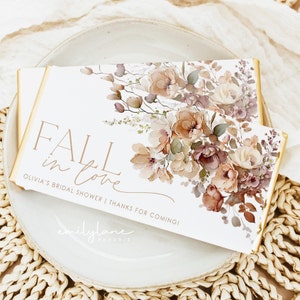 May include: Fall in love bridal shower favor chocolate bar wrapper with a watercolor floral design in shades of brown, pink, and green. The text reads "Fall in love" and "Olivia's Bridal Shower | Thanks for coming!" with the company name "emilylane paperie" at the bottom.