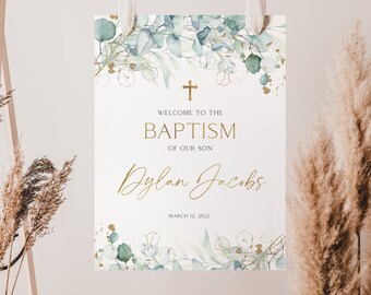 May include: A white sign with a gold cross at the top and a watercolor border of green leaves. The text on the sign reads "Welcome to the Baptism of our son Dylan Jacobs March 12, 2022".