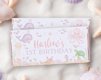 May include: A rectangular candy bar wrapper featuring a light purple and pink underwater theme with illustrations of a whale, octopus, jellyfish, crab, and sea turtle. The text reads Harlow's 1ST BIRTHDAY in a soft pink script and serif font.