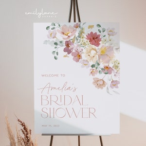 May include: A white welcome sign with a floral design in shades of pink, cream, and green. The text reads "Welcome to Amelia's Bridal Shower May 14, 2022".