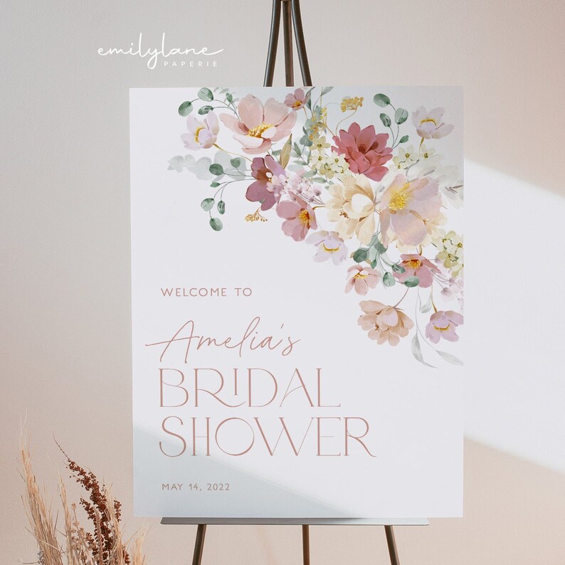 May include: A white welcome sign with a floral design in shades of pink, cream, and green. The text reads "Welcome to Amelia's Bridal Shower May 14, 2022".
