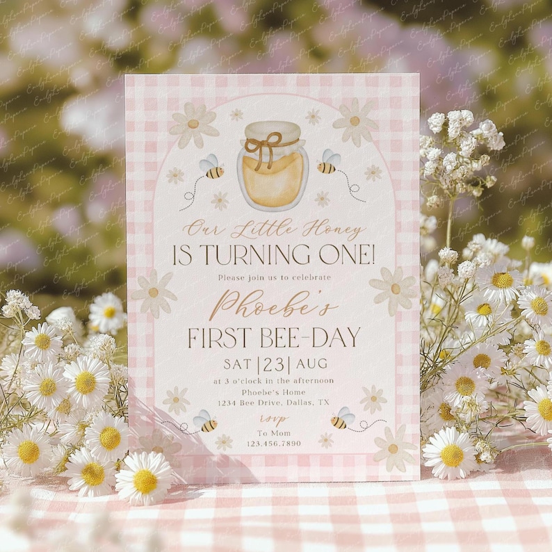 May include: A pink and white invitation card with the text "Our Little Honey Is Turning One!" and "Phoebe's First Bee-Day." The card features a honey jar illustration, bees, and daisy accents. The invitation includes date, time, and location details.