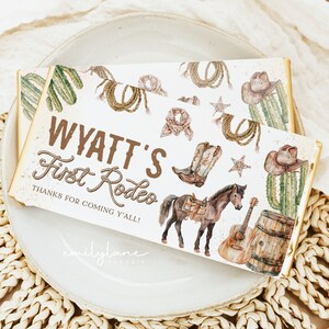 May include: A white chocolate bar wrapper with a western theme. It features a brown horse with a saddle, a guitar, a cowboy hat, and a cactus. The text on the wrapper reads "Wyatt's First Rodeo" and "Thanks for coming y'all!"