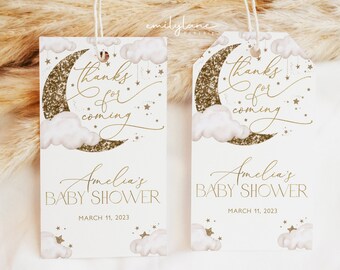 May include: Two white favor tags with gold glitter moons and clouds. The text on the tags reads "Thanks for coming Amelia's Baby Shower March 11, 2023".