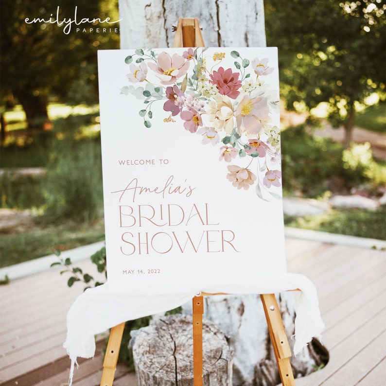 May include: A white welcome sign with pink floral illustrations and the text "Welcome to Amelia's Bridal Shower May 14, 2022".