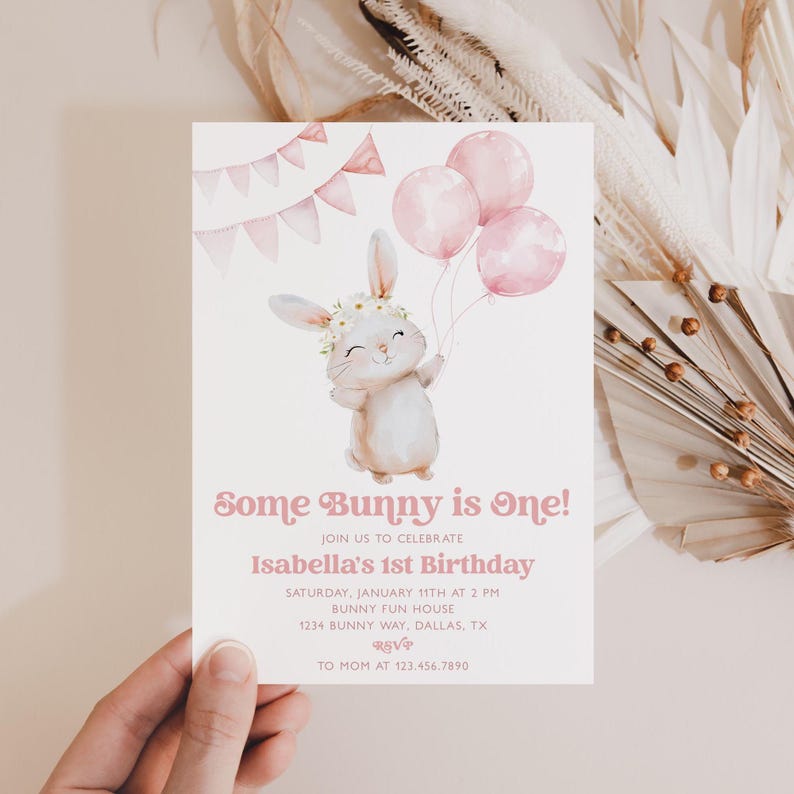 Op de afbeelding: Een roze en witte verjaardagsuitnodiging met een aquarel-illustratie van een konijn dat roze ballonnen vasthoudt. De tekst luidt "Some Bunny is One!" en bevat details voor Isabella's 1e verjaardag.