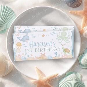 Puede incluir: Dos envoltorios rectangulares de barra de caramelo con un diseño de temática marina, con ballenas, un pulpo y el texto "1st Birthday de Harrison". Los envoltorios están sobre un plato blanco, rodeados de conchas marinas y una estrella de mar.