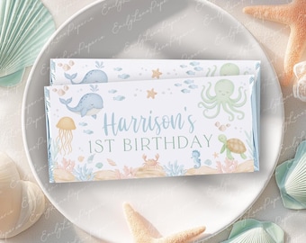 May include: Two rectangular candy bar wrappers with a sea-themed design, featuring whales, an octopus, and the text "Harrison's 1st Birthday." The wrappers are on a white plate, surrounded by seashells and a starfish.