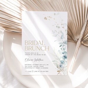 May include: A white invitation card with gold lettering for a bridal brunch. The card features a watercolor illustration of eucalyptus leaves and gold splatters. The text reads "Bridal Brunch", "Please join us in honor of the bride-to-be Olivia Wallace", "Saturday, October 30th at 2 PM", "Ida Claire, Dallas, Texas", "Registered at Target & Amazon", "RSVP to Jane at 123.456.7890".