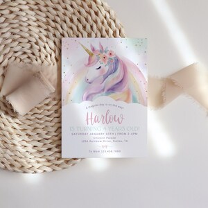 May include: A pastel-colored invitation card featuring a watercolor unicorn with a rainbow mane and floral crown. The card reads "Harlow is turning 4 years old!" and includes event details. The card is set on a woven placemat with a ribbon.