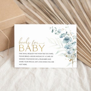 May include: A white card with gold lettering that says "books for baby". The card has a watercolor illustration of eucalyptus leaves in shades of blue and green with gold glitter accents. The text on the card reads: "One small request that won't be too hard. Please bring a book instead of a card. By signing your book we'll remember and share your special gift, even when you are not there."