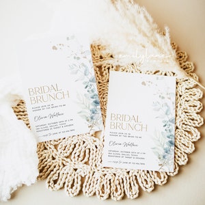 May include: Two white cards with gold lettering and blue watercolor floral designs. The text on the cards reads "Bridal Brunch" and includes the bride-to-be's name, date, time, and location. The cards are on a woven straw mat.