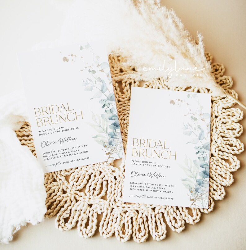 May include: Two white cards with gold lettering and blue watercolor floral designs. The text on the cards reads "Bridal Brunch" and includes the bride-to-be's name, date, time, and location. The cards are on a woven straw mat.