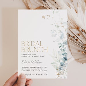 May include: A white invitation card with gold lettering announcing a bridal brunch for Olivia Wallace. The card features a watercolor illustration of eucalyptus leaves and gold accents.