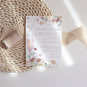 May include: A floral-themed bridal shower invitation with a glass of pink prosecco. The invitation reads "Petals, Pearls & Prosecco" and includes the bride-to-be's name, date, time, and location. The invitation also includes the text "RSVP to Jane at [phone number]" and "Registered at Amazon & Target".