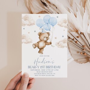 May include: A blue and white invitation for a 1st birthday party with a teddy bear holding blue balloons. The invitation says "Join us for Hudson's Bear-Y 1st Birthday, Saturday, May 17th at 1 PM, Bear Creation Shop, 1234 Bear Drive, Dallas, TX, RSVP to Mom 123.456.7890".